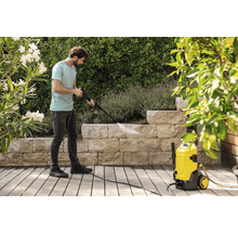 Een man reinigt een stenen muur in een tuin met een gele hogedrukreiniger op een houten terras omgeven door planten.