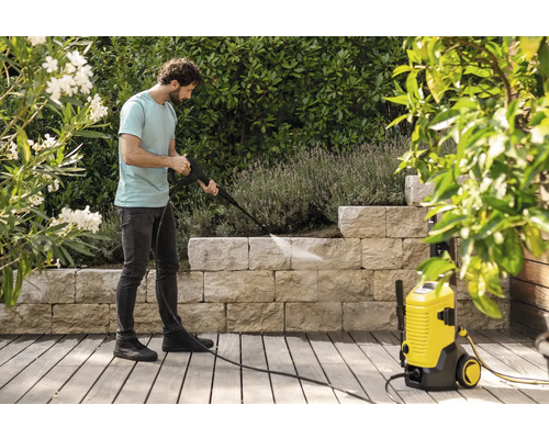 Een man reinigt een stenen muur in een tuin met een gele hogedrukreiniger op een houten terras omgeven door planten.
