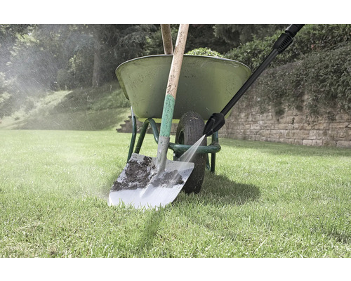 Een hogedrukreiniger reinigt een vuile schep voor een groene kruiwagen op een gazon.