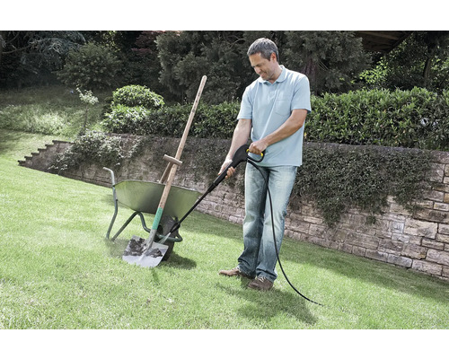 Een man reinigt een vuile schep in een kruiwagen met een hogedrukreiniger op een gazon in de tuin.