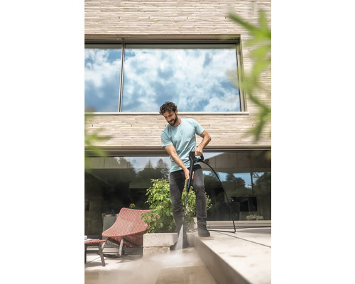 Een man reinigt een licht stenen terras voor een modern huis met een hogedrukreiniger.