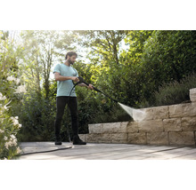 Een man reinigt een natuurstenen muur op een houten terras in de tuin met een hogedrukreiniger in een lichte, georganiseerde omgeving.