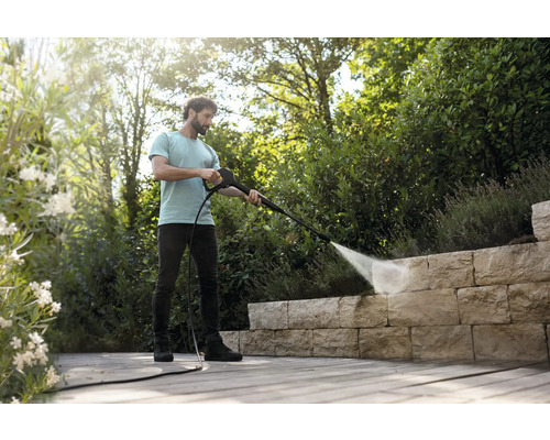 Een man reinigt een natuurstenen muur op een houten terras in de tuin met een hogedrukreiniger in een lichte, georganiseerde omgeving.