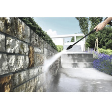 Reinigen van een natuurstenen muur in een lichte tuin met een hogedrukreiniger, naast stenen trappen en planten.