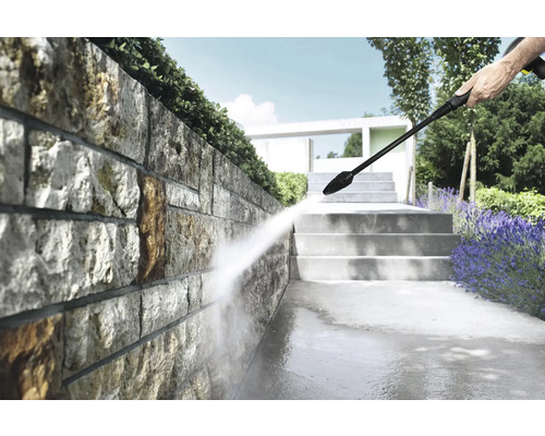 Reinigen van een natuurstenen muur in een lichte tuin met een hogedrukreiniger, naast stenen trappen en planten.