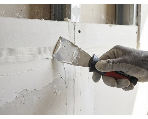 Hand met werkhandschoen brengt met een Rautner plamuurmes vulmiddel aan op een voegband op een gipsplaten wand.