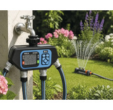 Digitale besproeiingscomputer van for.q op een buitenkraan met aangesloten tuinslangen en een actieve gazonsproeier in de tuin.