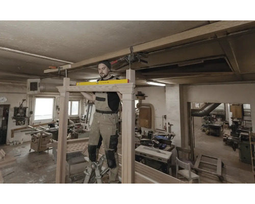 Vakman in een werkplaats gebruikt een waterpas om een houten frame uit te lijnen terwijl hij op een ladder staat.