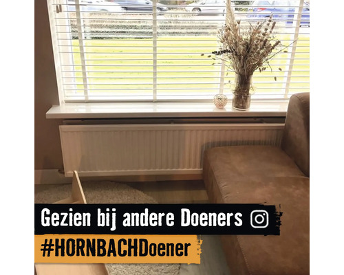 Paneelradiator onder een raam met witte jaloezieën en een vaas op de vensterbank naast een bank in een lichte woonkamer.