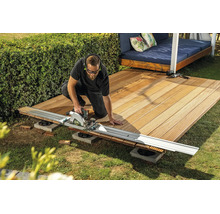 Een vakman zaagt houten terrasplanken met een cirkelzaag en geleiderail in de tuin.