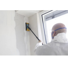 Persoon in beschermende kleding schuurt een gestuukte muur met een handschuurmachine op een telescoopsteel.