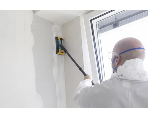 Persoon in beschermende kleding schuurt een gestuukte muur met een handschuurmachine op een telescoopsteel.