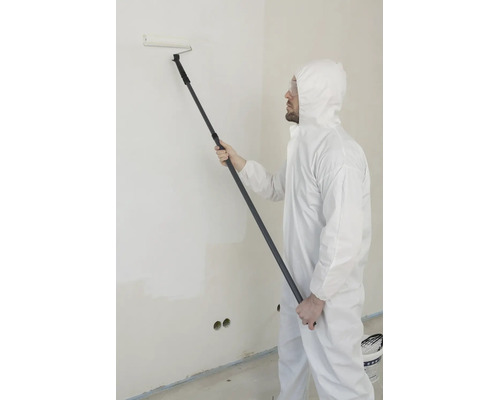 Man in wit beschermend pak en veiligheidsbril verft een muur met een verfroller aan een telescoopsteel.
