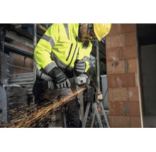 Vakman met Metabo accu-haakse slijper snijdt metaal op een bouwplaats, vonkenregen, beschermende kleding en helm.
