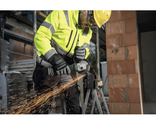 Vakman met Metabo accu-haakse slijper snijdt metaal op een bouwplaats, vonkenregen, beschermende kleding en helm.