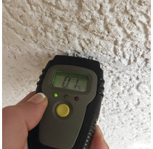 Een digitale vochtmeter wordt tegen een gestructureerde muur gehouden en toont een waarde van nul komma één procent.