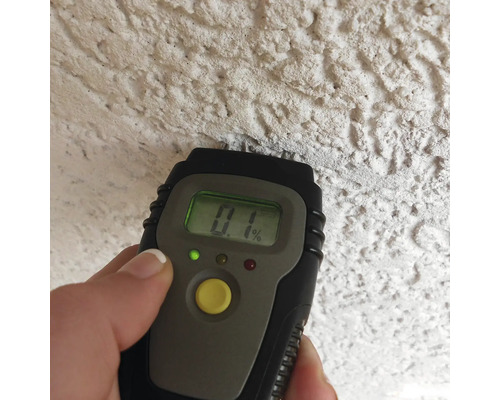 Een digitale vochtmeter wordt tegen een gestructureerde muur gehouden en toont een waarde van nul komma één procent.