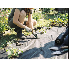 Een persoon legt worteldoek in de tuin en zet het vast met grondpennen en een hamer.