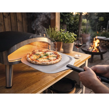 Vers gebakken pizza wordt met een pizzaschep met Tenneker Logo uit een draagbare pizzaoven op een houten tafel in de tuin gehaald.