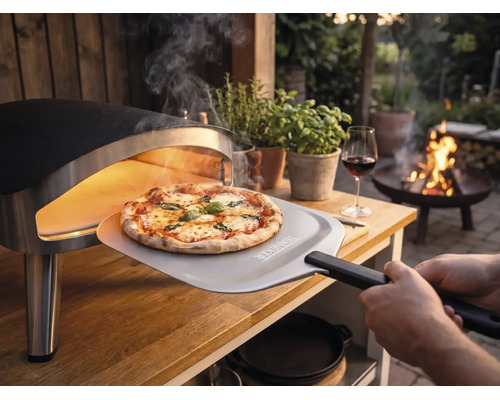Vers gebakken pizza wordt met een pizzaschep met Tenneker Logo uit een draagbare pizzaoven op een houten tafel in de tuin gehaald.