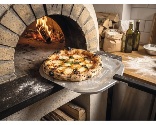 Versgebakken pizza op een pizzaschep met Tenneker Logo voor een brandende steenoven in een rustieke keuken.