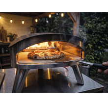 Roestvrijstalen pizzaoven voor buiten met brandende vlammen en een pizza op een pizzasteen die met een pizzaschep wordt verplaatst.