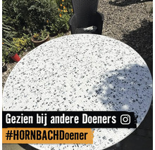 Ronde tuintafel met een mozaïek tafelblad van tegels met zwart-wit motieven in een lichte tuin naast een stoel. Instagram Logo.