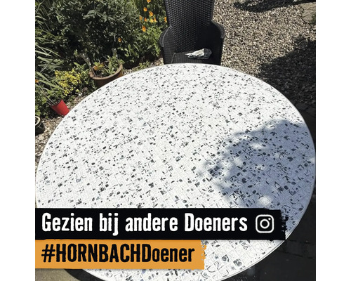 Ronde tuintafel met een mozaïek tafelblad van tegels met zwart-wit motieven in een lichte tuin naast een stoel. Instagram Logo.