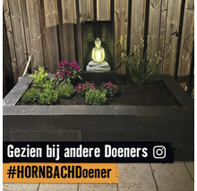 Verhoogde plantenbak van donkere stapelblokken met diverse planten en een verlicht Boeddha waterornament voor een houten schutting.