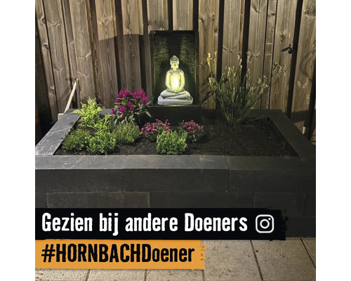 Verhoogde plantenbak van donkere stapelblokken met diverse planten en een verlicht Boeddha waterornament voor een houten schutting.