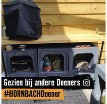 Zelfgebouwde buitenkeuken van modulaire stenen blokken en houten werkblad voor een houten wand. Er ligt een kat in een van de blokken.