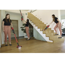 Een persoon gebruikt een rode snoerloze stofzuiger in een lichte kamer voor het reinigen van vloeren, houten trappen en hoge hoeken.