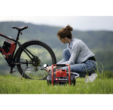 Een vrouw pompt de achterband van een fiets op met een Einhell accu-compressor op een grasveld.