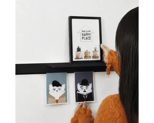 Iemand decoreert een zwarte wandplank met een fotolijst en twee kattenkaarten aan een witte muur.