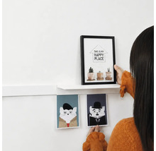 Een persoon decoreert een muur met een witte wandplank, een fotolijst met plantenmotief en twee kattenafbeeldingen als wanddecoratie.