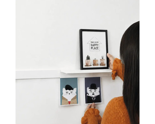 Een persoon decoreert een muur met een witte wandplank, een fotolijst met plantenmotief en twee kattenafbeeldingen als wanddecoratie.