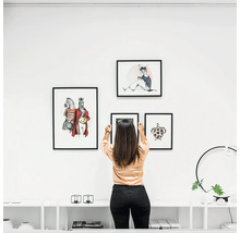 Vrouw hangt fotolijsten met dierenafbeeldingen aan een witte muur boven een dressoir in een modern, licht interieur.