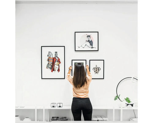 Vrouw hangt fotolijsten met dierenafbeeldingen aan een witte muur boven een dressoir in een modern, licht interieur.