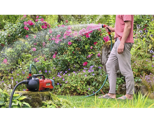 Einhell accu tuinpomp Aquinna bij het besproeien van de tuin met een tuinslang en een spuitpistool in een beplante tuin.