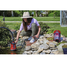 Vrouw bedient een Einhell accu-schoonwaterpomp bij een lichte tuinvijver met stenen en planten.