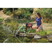 Een vrouw bedient een rode Einhell tuinpomp bij een vijver voor tuinbewatering met een sproeier op de achtergrond.