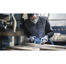 Vakman schuurt een houten plank met een Bosch bandschuurmachine in een werkplaats met beschermende kleding en een stofmasker.