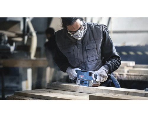Vakman schuurt een houten plank met een Bosch bandschuurmachine in een werkplaats met beschermende kleding en een stofmasker.