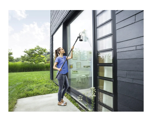Vrouw reinigt een hoog glazen pui aan een modern huis met een accu raamwisser met verlengset.