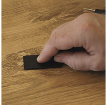 Een hand gebruikt een zwarte schraper om reparatiewas op een houten vloeroppervlak aan te brengen om een beschadiging te herstellen.
