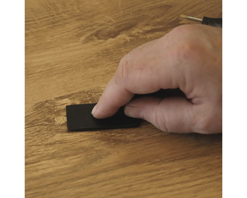 Een hand gebruikt een zwarte schraper om reparatiewas op een houten vloeroppervlak aan te brengen om een beschadiging te herstellen.