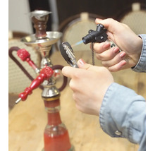 Een hand steekt shishakool aan met een gasbrander, op de achtergrond staat een waterpijp op een tafel.