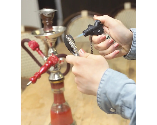 Een hand steekt shishakool aan met een gasbrander, op de achtergrond staat een waterpijp op een tafel.