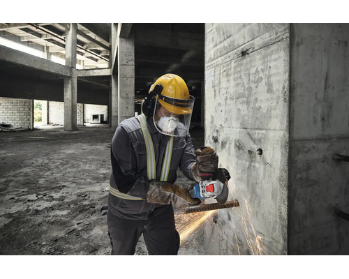 Een vakman met beschermende kleding gebruikt een haakse slijper om een metalen staaf bij een betonnen muur op een bouwplaats door te slijpen.