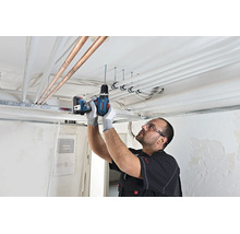 Vakman boort met Bosch accuschroefmachine in een betonnen plafond met leidingen op een bouwplaats.